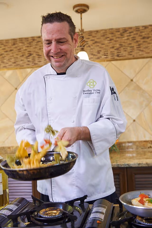 A male chef wearing a white chef coat with the name Bradley Zorian and title Executive Chef is cooking pasta in a frying pan over a gas stove in a kitchen with tiled walls and granite countertops.