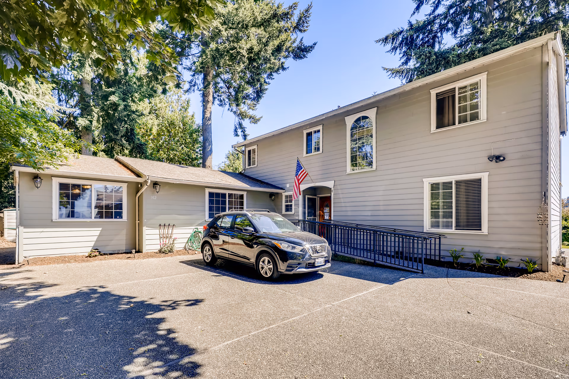 Exterior view of a two-story gray building with white trim, an American flag near the entrance, a black car parked in front, and surrounded by tall trees under a clear blue sky.