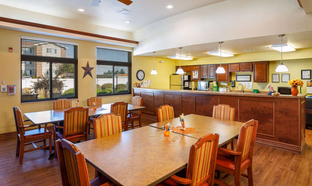 A bright dining area in an assisted living facility with several wooden tables and striped cushioned chairs. The room features large windows with blinds, a wall clock, and a kitchen area with wooden cabinets, a refrigerator, microwave, and pendant lights hanging over the counter. Seasonal decorations are placed on the tables and counter.