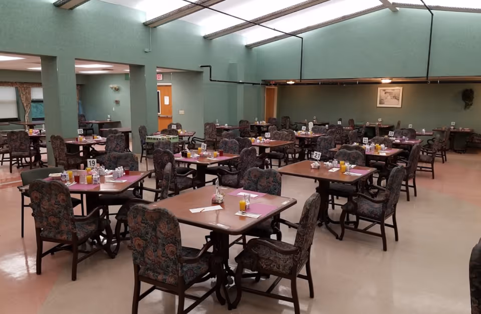 A large dining room with multiple tables and chairs arranged neatly. Each table has placemats, glasses of orange juice, and condiments. The room has green walls and a ceiling with fluorescent lighting. There are table number signs on each table.