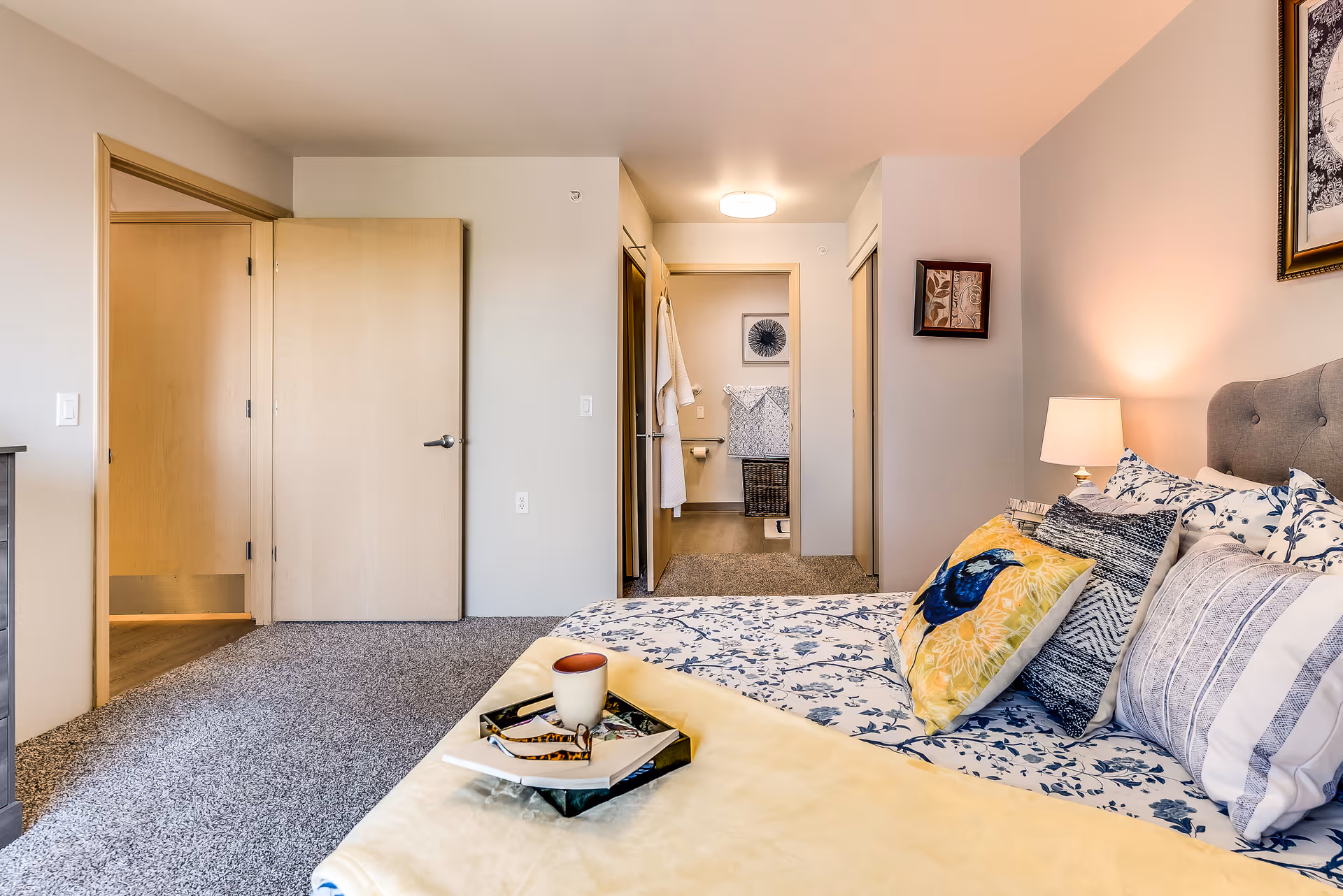 Bright furnished bedroom with a made bed, decorative pillows, bedside lamp, and an open doorway leading to a bathroom.