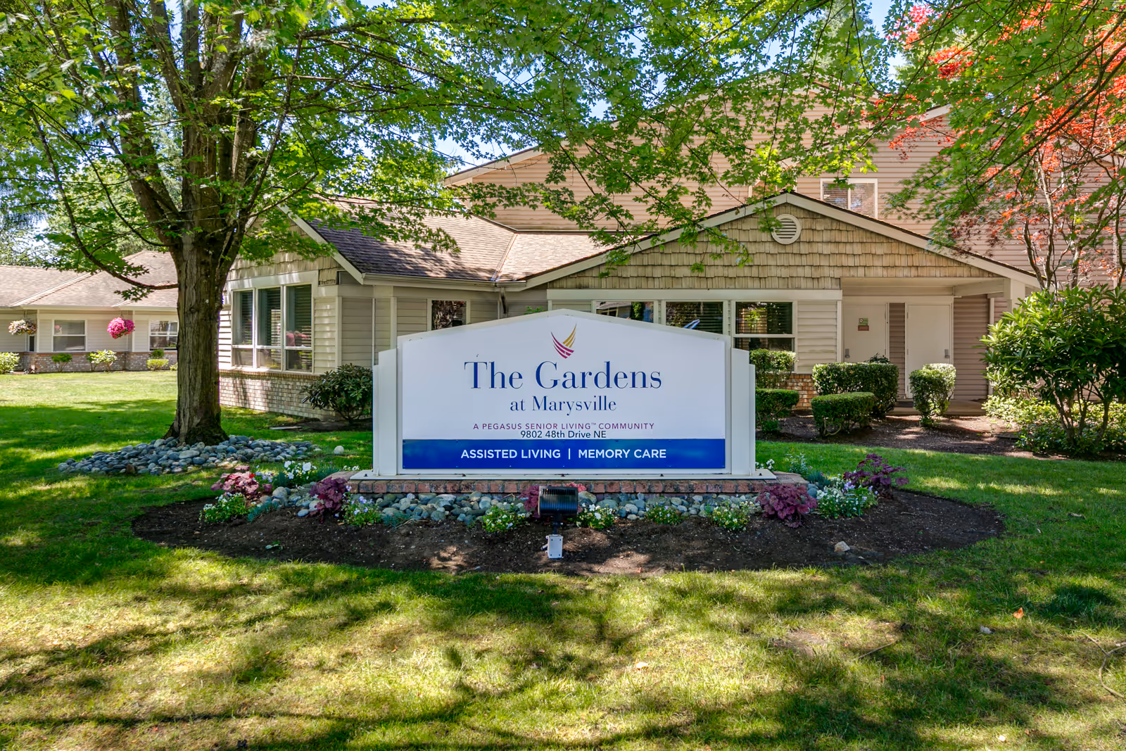 The exterior of The Gardens at Marysville senior living facility with a large sign in front that reads 'The Gardens at Marysville, A Pegasus Senior Living Community, Assisted Living | Memory Care'. The building is surrounded by green grass, trees, and shrubs under a sunny sky.