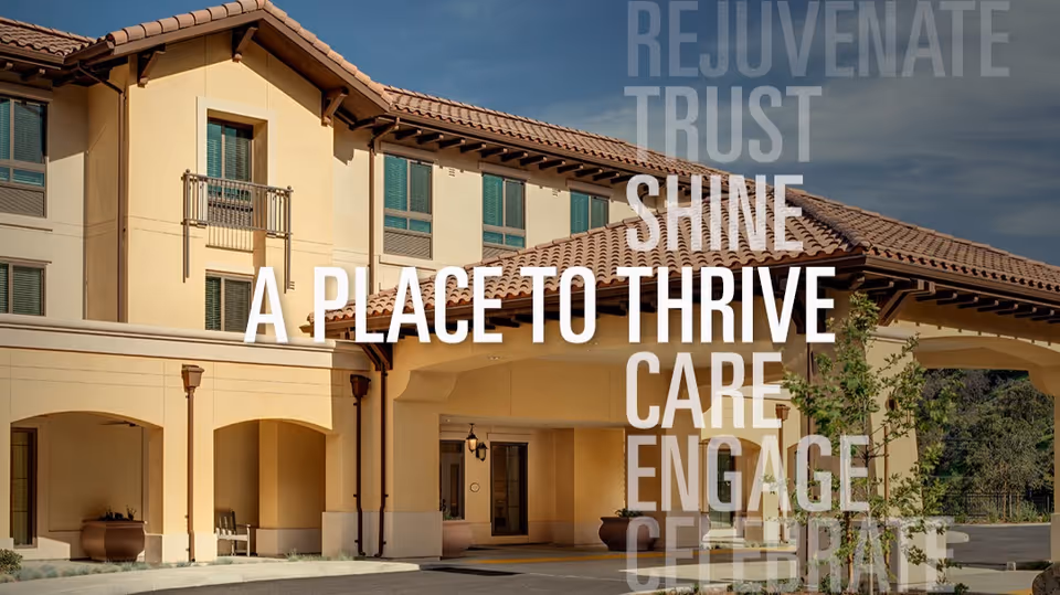 Exterior view of a senior living facility building with beige walls and a tiled roof under a partly cloudy sky. The entrance is covered with an archway, and there are potted plants near the entrance. Overlaid text reads: 'REJUVENATE TRUST SHINE A PLACE TO THRIVE CARE ENGAGE CELEBRATE'.