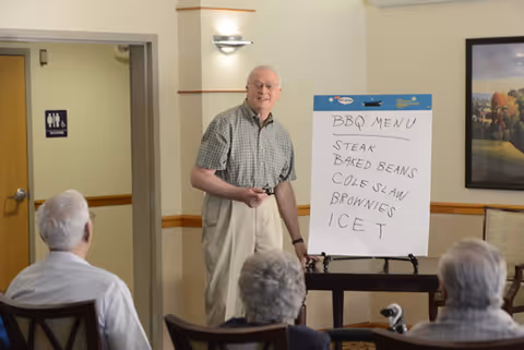 An elderly man standing and pointing at a whiteboard with a BBQ menu written on it, addressing a small group of seated elderly people in a room with beige walls and a framed picture on the wall.