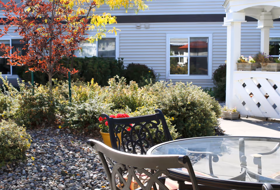 Sunlit courtyard with a glass-top patio table and chairs in front of the senior living building's exterior and landscaping.