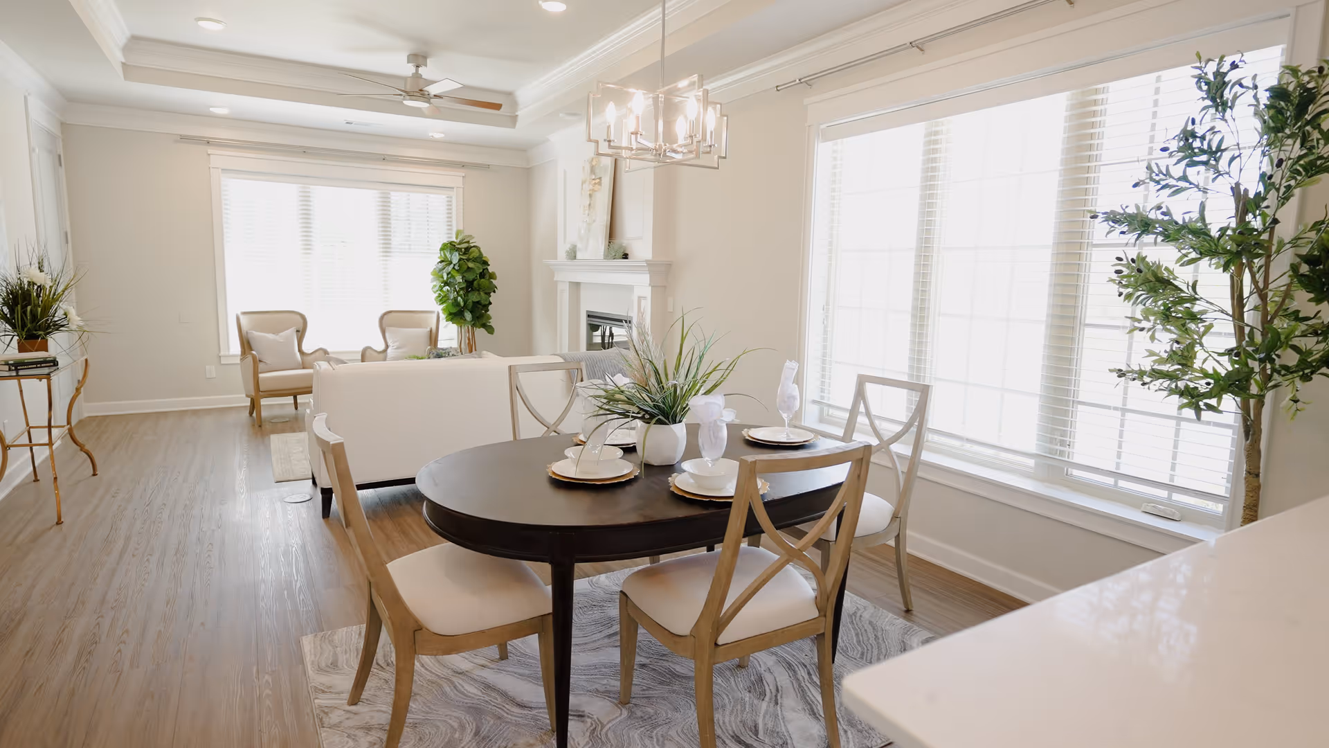 Bright and airy living and dining area with a dark wooden oval dining table set with four chairs, white cushioned seats, and table settings. The room features large windows with white blinds, light-colored walls, a ceiling fan, a modern chandelier, a white sofa, two beige armchairs, a fireplace, and several green plants.