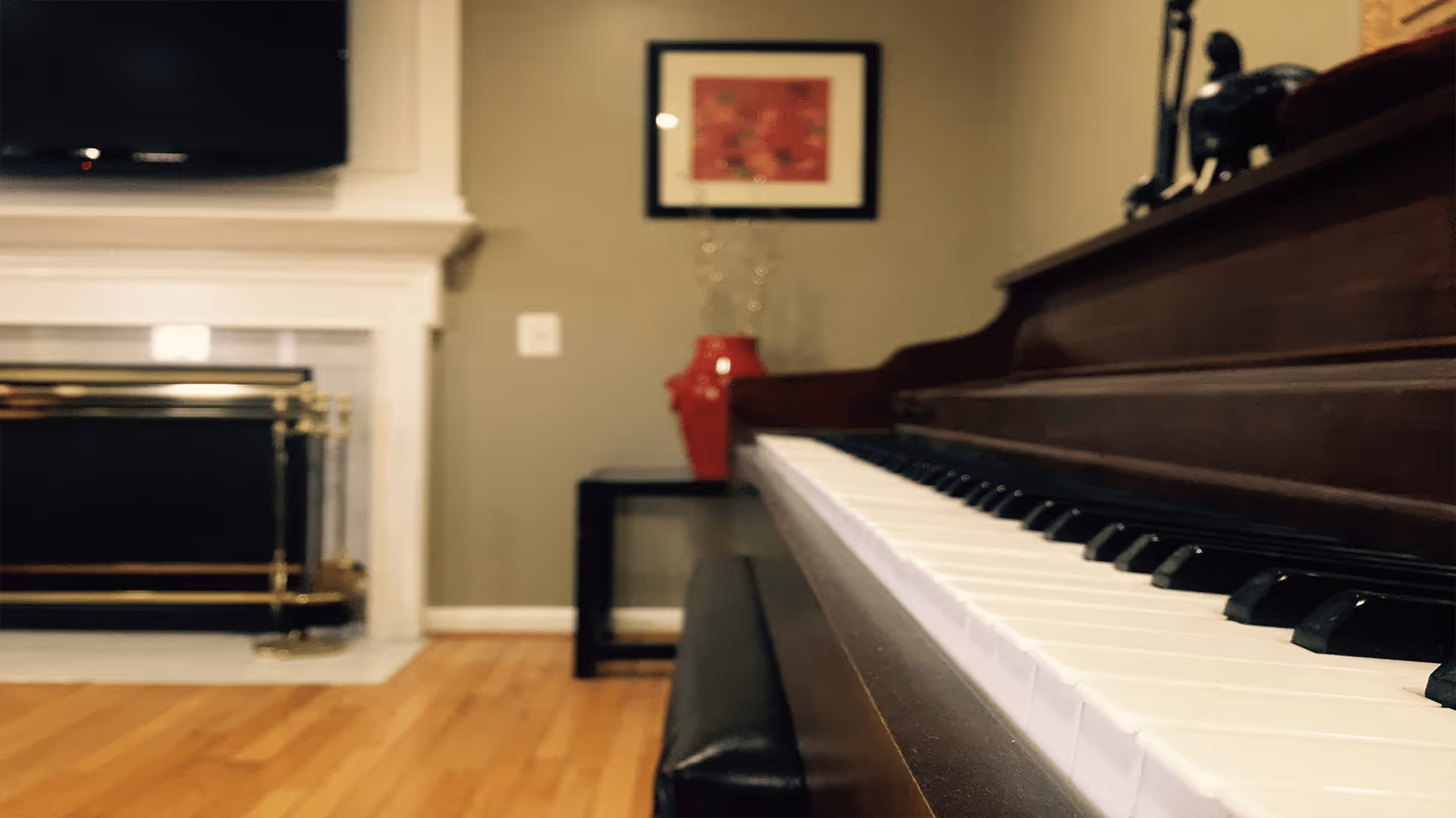 Close-up view of a piano keyboard in a cozy living room with a hardwood floor, a fireplace with a brass screen, a small black side table holding a red vase, and a framed picture on the wall.