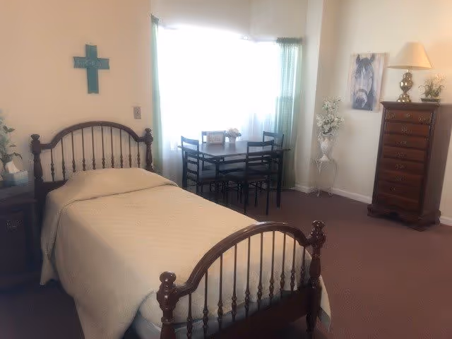 A cozy bedroom with a single bed featuring a wooden headboard and footboard, covered with a beige blanket. There is a small wooden nightstand with a tissue box on the left side of the bed. A green cross hangs on the wall above the bed. In the background, a window with sheer white curtains and green drapes lets in natural light. A small dining table with four chairs is placed near the window. On the right side, there is a tall wooden dresser with a lamp and a framed picture of a horse above it. A decorative plant stands next to the dresser.