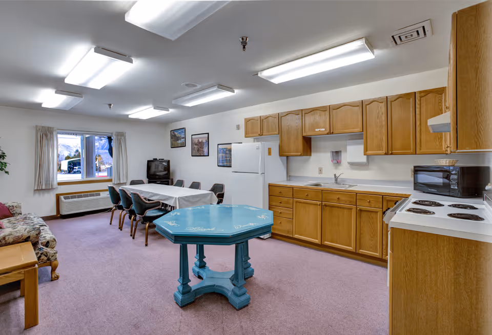 Interior view of a senior living facility common area featuring a kitchen with wooden cabinets, a white refrigerator, a microwave, and an electric stove. There is a blue octagonal table in the center, a dining table with chairs covered with a white tablecloth near a window, and a floral-patterned couch with a wooden side table. The room has carpeted flooring and fluorescent ceiling lights.