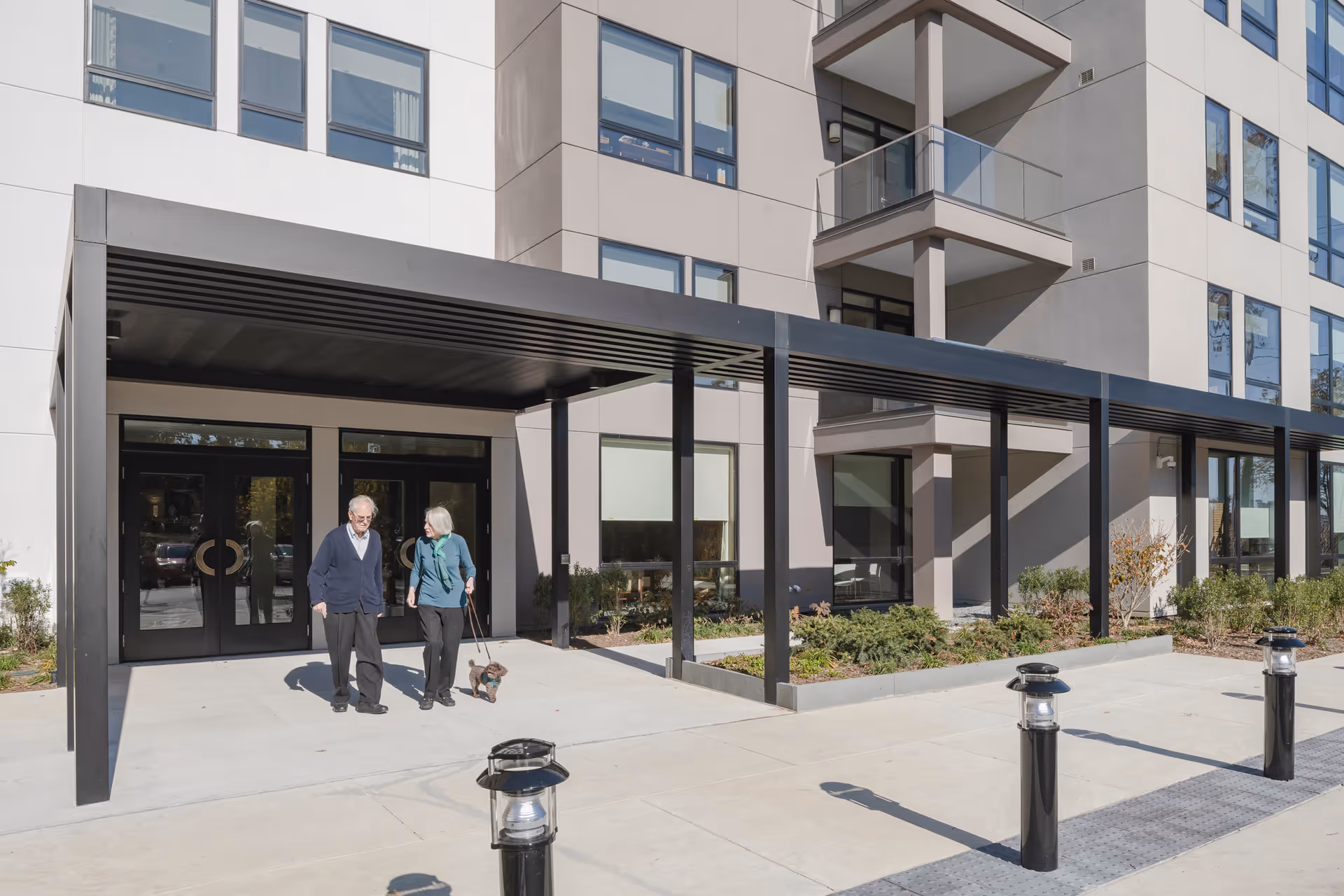 Two elderly people walking a small dog outside the entrance of a modern multi-story senior living facility with large windows and a covered walkway.