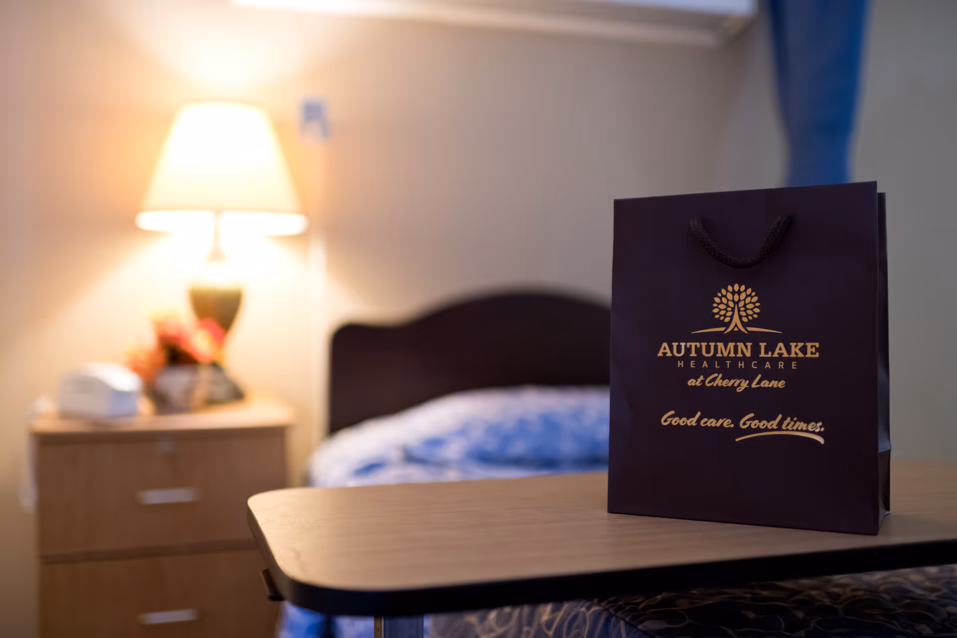 A bedside table with a lamp and a telephone next to a bed with blue patterned bedding. In the foreground, there is a dark-colored gift bag with the text 'Autumn Lake Healthcare at Cherry Lane Good care. Good times.'