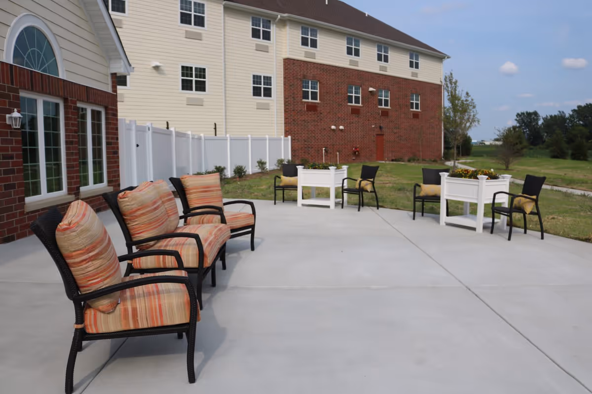 Outdoor patio with cushioned chairs and white planters in front of a multi-story senior housing building.