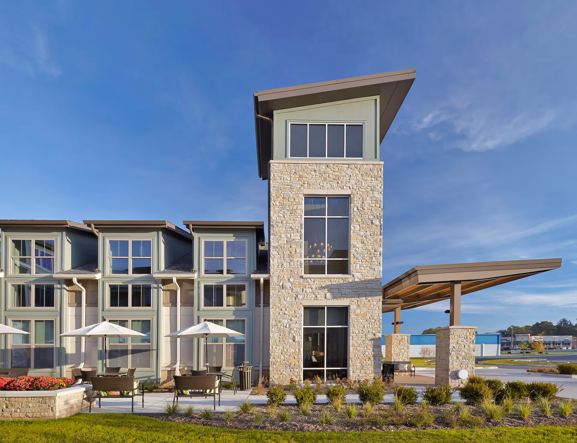 Exterior view of Wellbrooke of South Bend facility showing a modern building with large windows, stone and green siding, outdoor seating with umbrellas, landscaped bushes, and a clear blue sky.