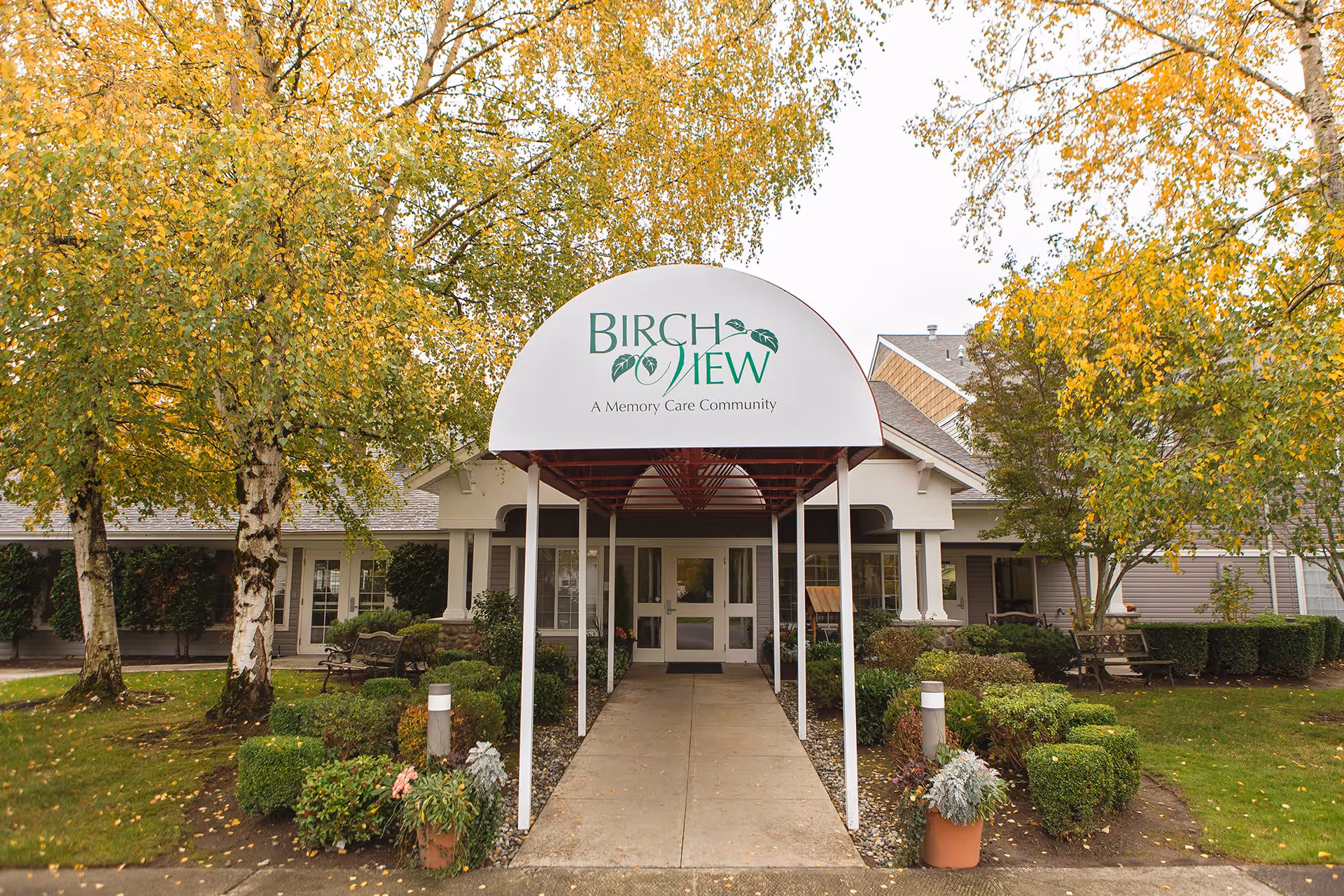 Entrance to Birchview Memory Care community with a covered walkway, surrounded by green and yellow-leaved trees and well-maintained bushes and plants.