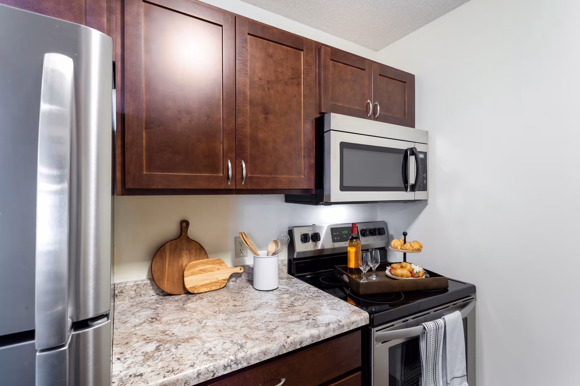 A modern kitchen corner featuring dark wooden cabinets, a stainless steel refrigerator, a stainless steel microwave mounted above a black electric stove with an oven. On the countertop, there are two wooden cutting boards, a white utensil holder with wooden spoons, and a tray on the stove holding a bottle of wine, two wine glasses, and a tiered plate with croissants and strawberries. A white towel hangs on the oven handle.