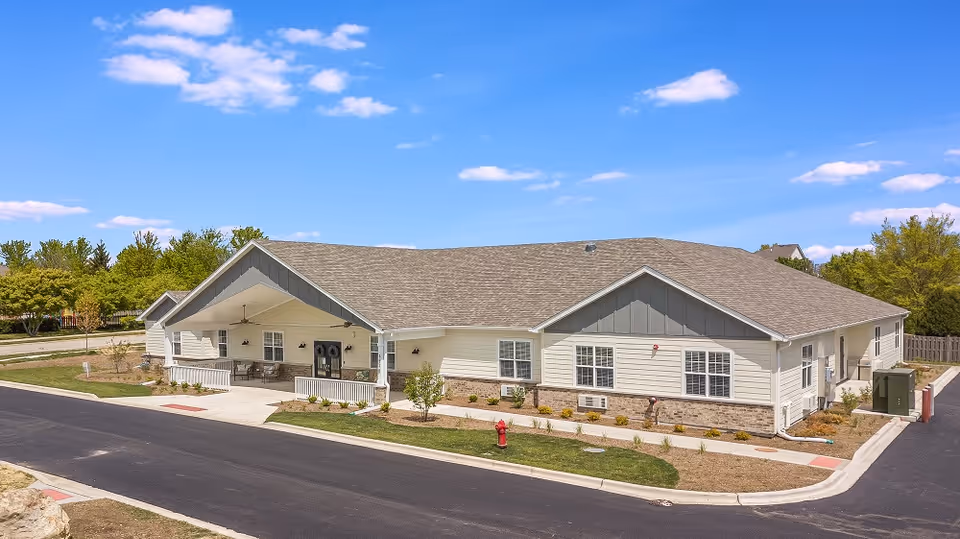 Exterior view of a single-story senior living facility building with a covered entrance, beige siding, and a gray shingled roof under a blue sky with scattered clouds. The building is surrounded by a paved road, small landscaped areas with grass and shrubs, and trees in the background.