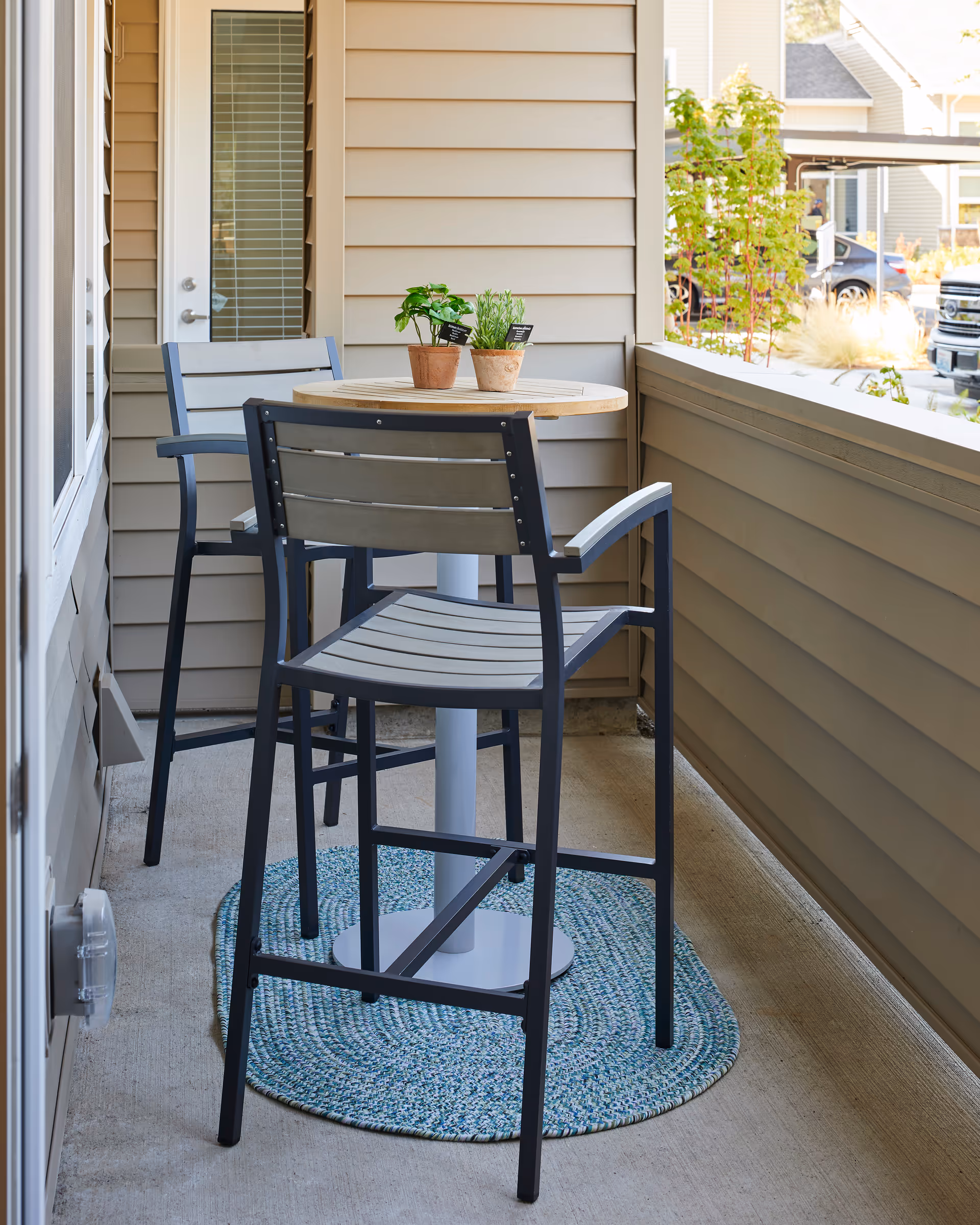 Small covered balcony with a round bistro table, two tall chairs, two potted plants, and a woven rug.
