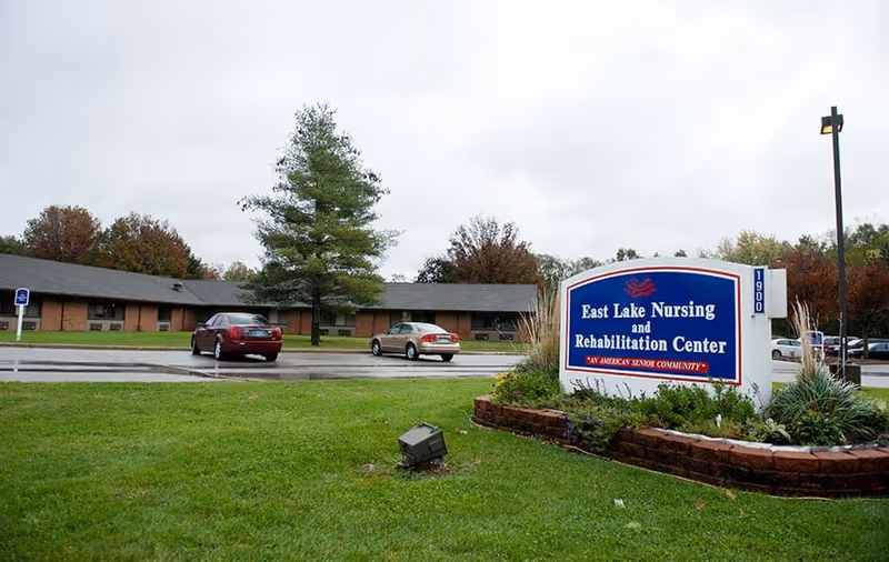 Front lawn and parking area of East Lake Nursing and Rehabilitation Center with a large blue entrance sign and a single-story brick building behind it.