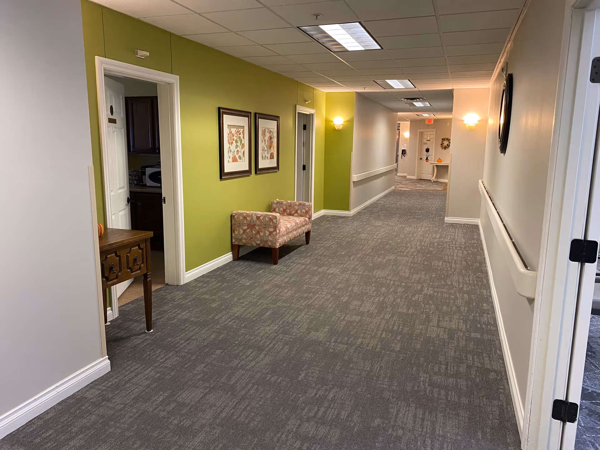 Carpeted interior hallway in an assisted living facility with a green accent wall, framed artwork, a patterned chair and handrails.