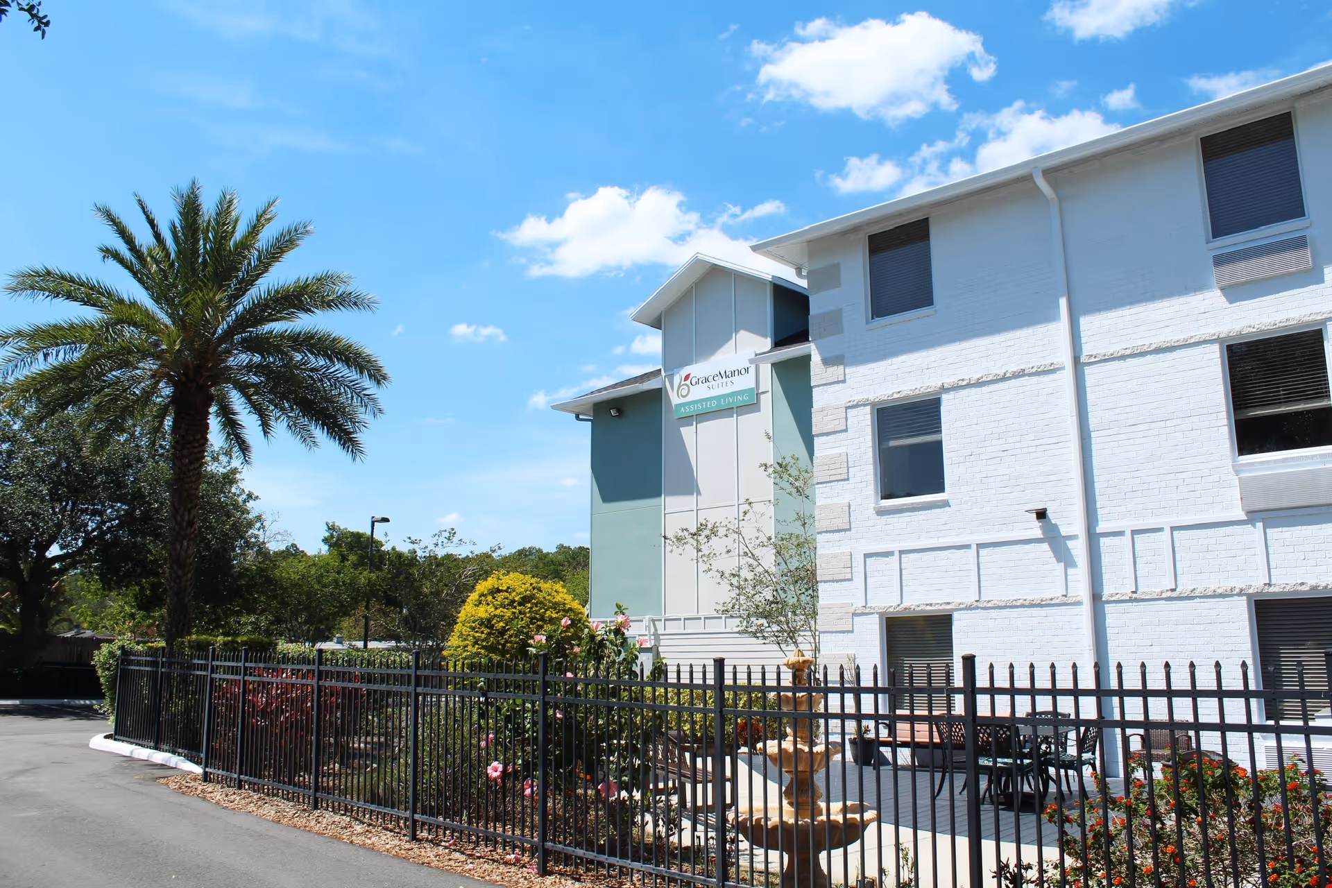 Exterior front of Grace Manor Suites assisted living building with a palm tree, wrought-iron fence, and courtyard fountain.