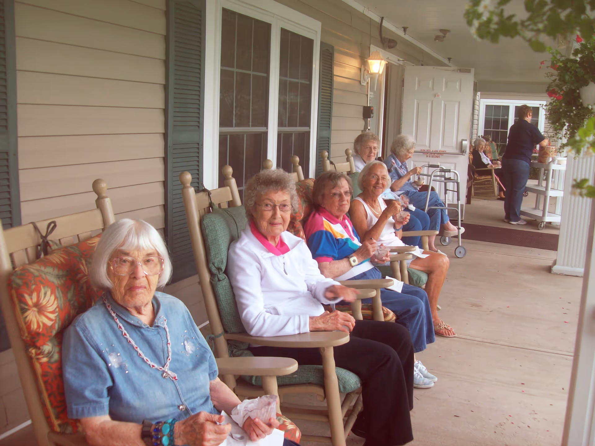 Several elderly women sitting in rocking chairs on a covered porch of a senior living facility, some eating ice cream, with a staff member standing near a cart in the background.