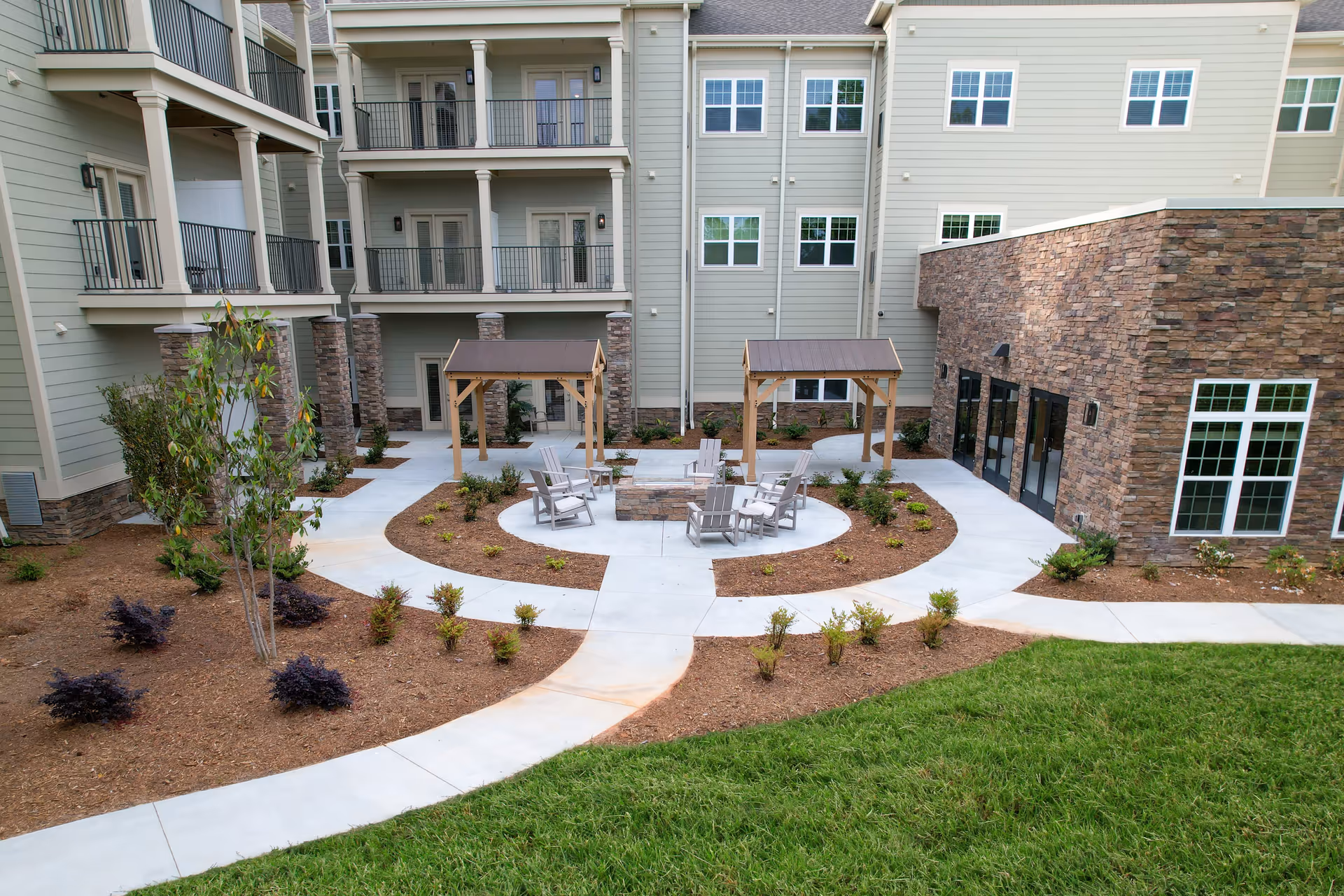 Outdoor courtyard area of a senior living facility with a circular concrete pathway surrounding a fire pit with six Adirondack chairs. Two wooden pergolas with brown roofs are positioned near the fire pit. The courtyard is landscaped with small shrubs and plants, and the building exterior features beige siding with stone accents and multiple windows and balconies.