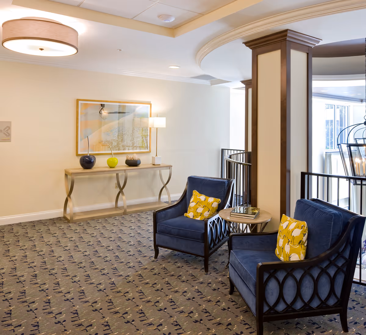 A cozy seating area in a senior living facility with two blue armchairs featuring yellow patterned pillows, a small round wooden side table with books, a decorative console table with three ornamental vases and a lamp, a framed abstract artwork on the wall, and a patterned carpeted floor under soft ceiling lighting.