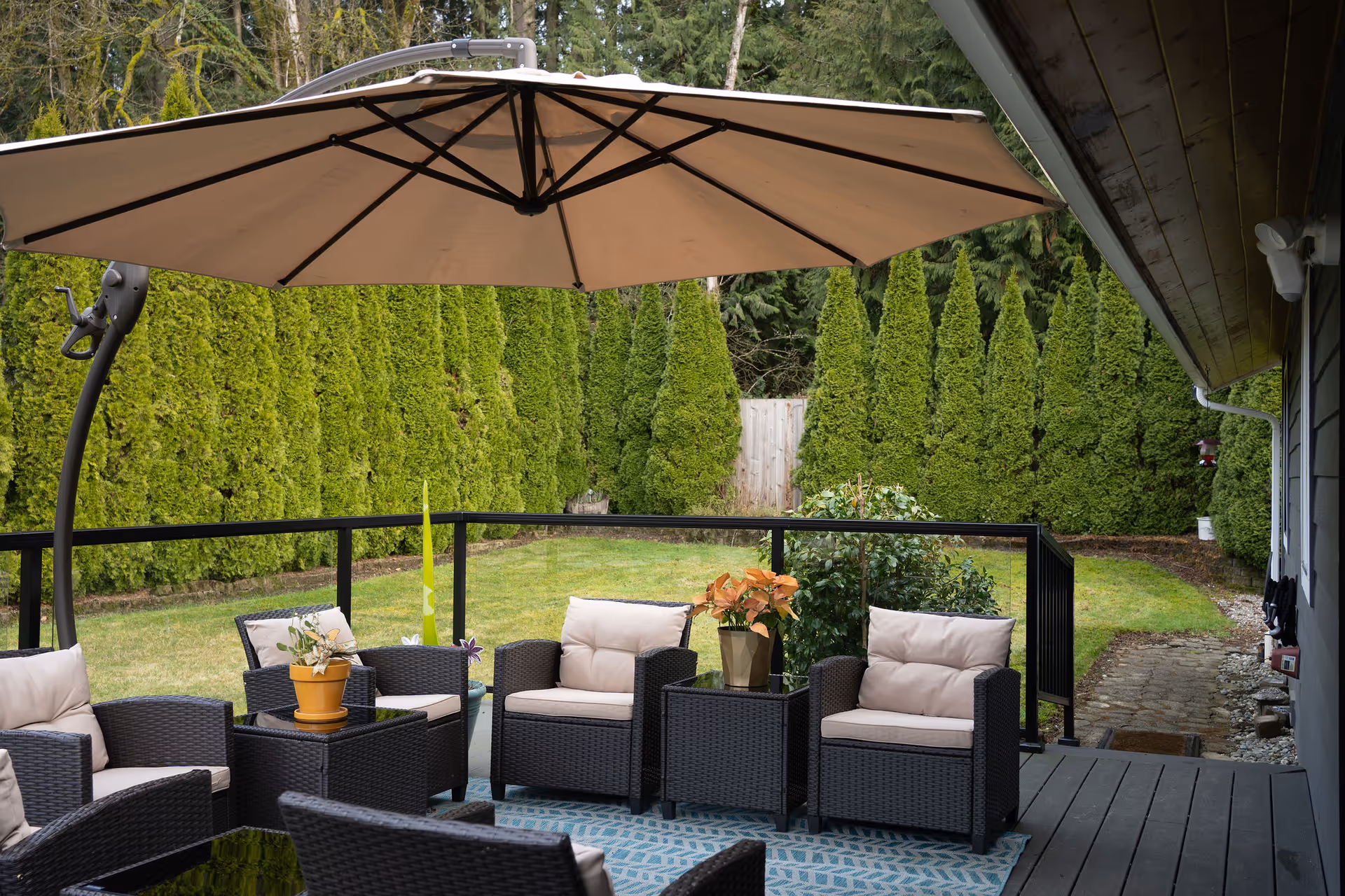 Outdoor patio area with cushioned wicker chairs and a large beige umbrella. The patio overlooks a green lawn bordered by tall, dense evergreen trees and a wooden fence in the background. There are potted plants on the tables and a patterned outdoor rug on the deck floor.