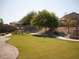 A landscaped outdoor area with a green lawn, a paved walkway curving through the grass, a tree providing shade, a gazebo structure, and a multi-tiered water fountain. There are some bushes and palm trees near a beige wall and a building in the background under a clear sky.