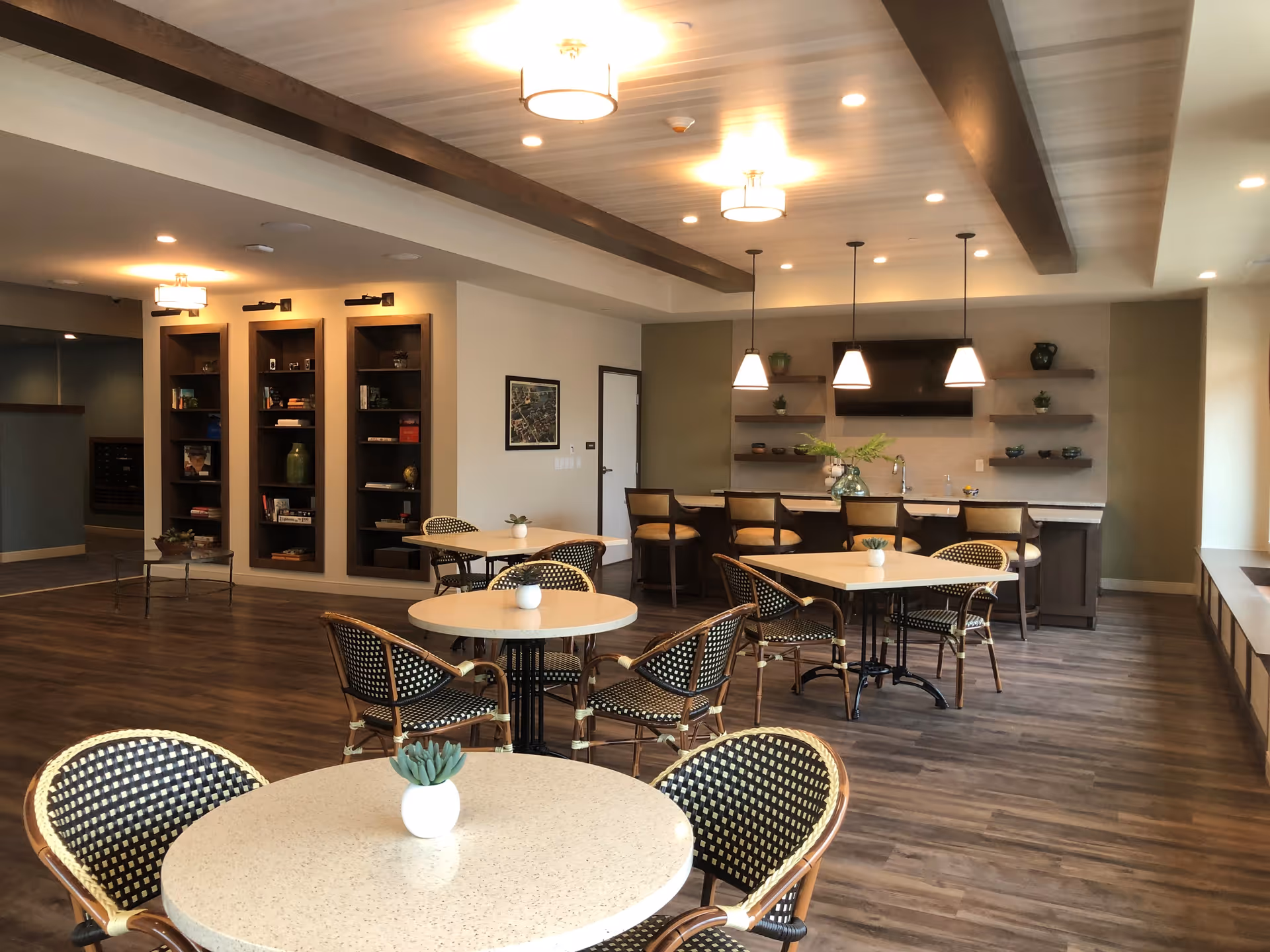 Communal dining area with round tables and wicker chairs facing a counter and shelves under pendant lighting.