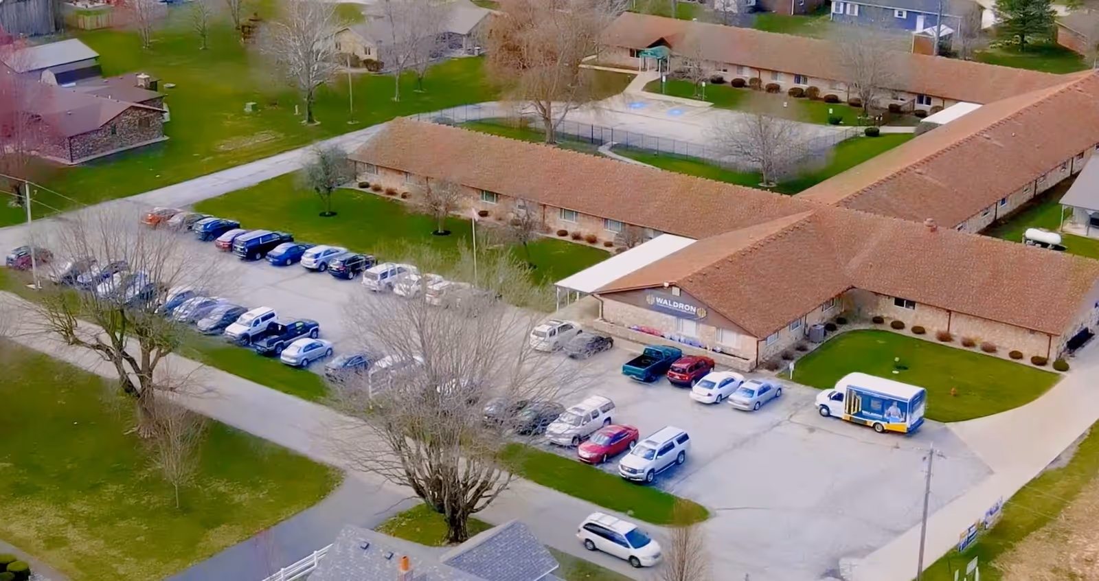 Aerial view of the Waldron Rehabilitation and Healthcare Center showing an L-shaped brick building, parking lot with cars, and surrounding lawns.