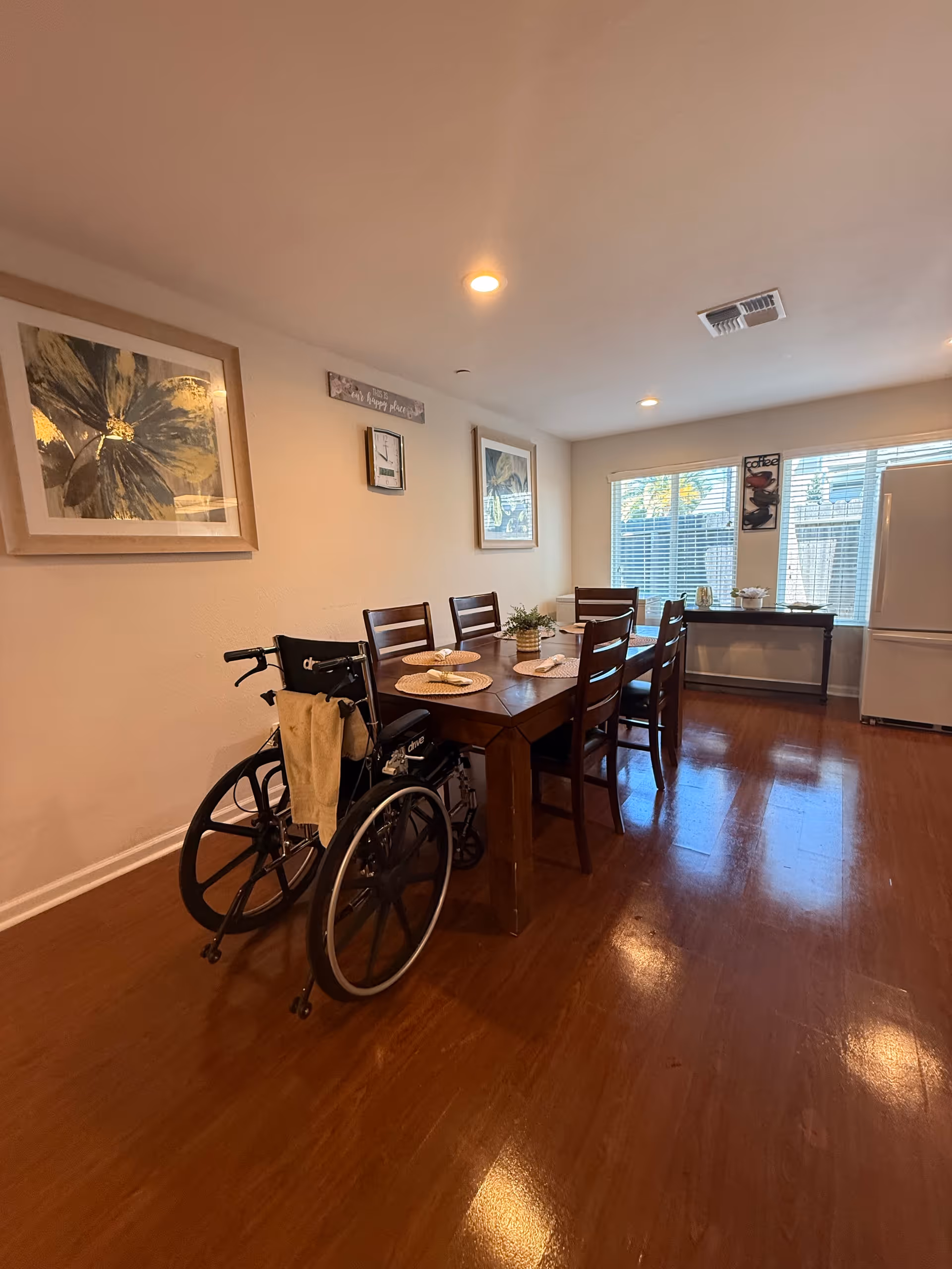 Dining room with a wooden table and chairs, a wheelchair beside the table, wall art on the walls, and large windows letting in light.