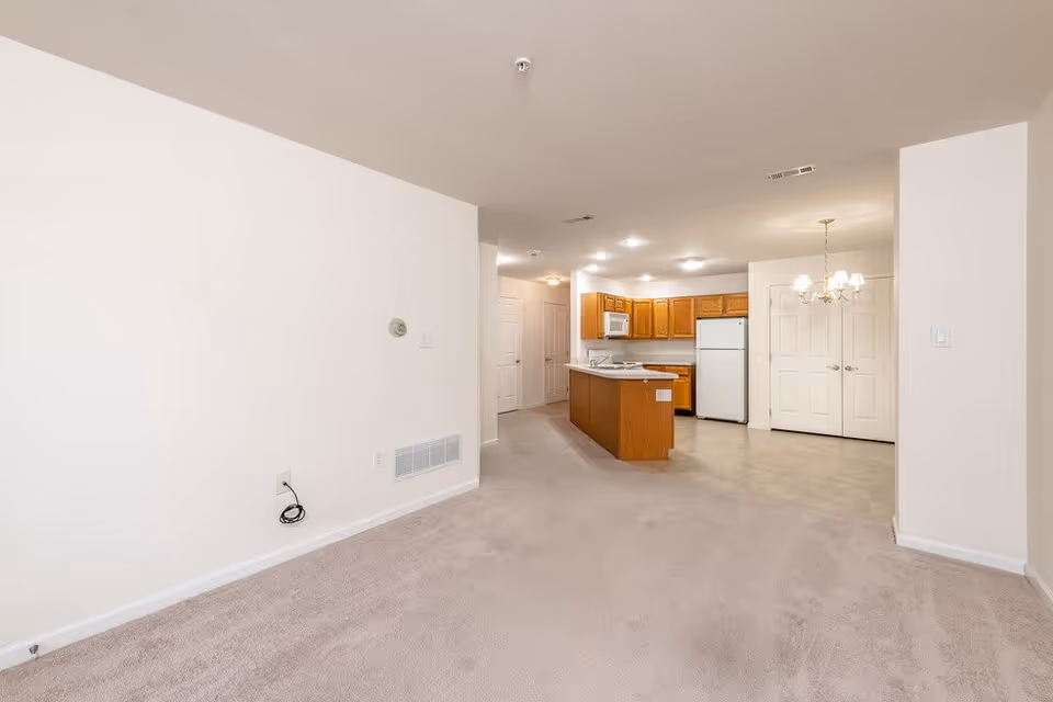 Interior view of an empty apartment showing a carpeted living area leading into a kitchen with wooden cabinets, white appliances including a refrigerator and microwave, and a small island. There is a chandelier hanging over a tiled dining area near double doors.