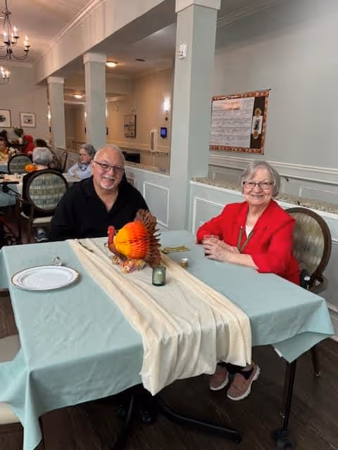 Two elderly individuals sitting at a dining table in a well-lit room with light-colored walls and decorative molding. The table is covered with a light blue tablecloth and a cream-colored runner, with a decorative turkey centerpiece and a small candle. Other people are visible seated at tables in the background.