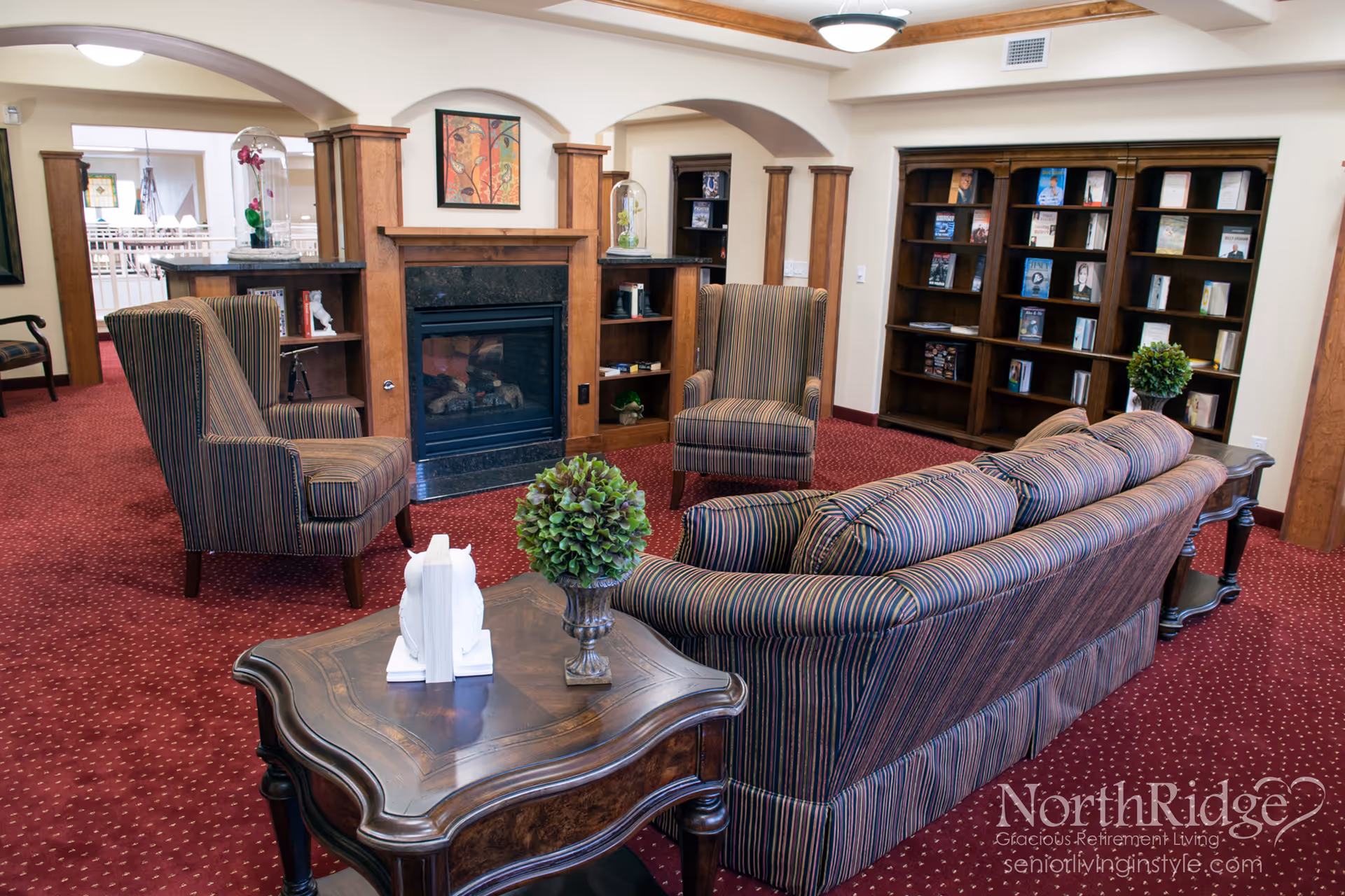 A cozy living room area in a retirement facility featuring a striped sofa and two matching armchairs arranged around a fireplace with wooden built-in shelves on either side. A wooden coffee table with a small green plant and white owl bookends is in the foreground. A large bookshelf filled with books is against the wall, and the room has red carpet and cream-colored walls with wooden trim.