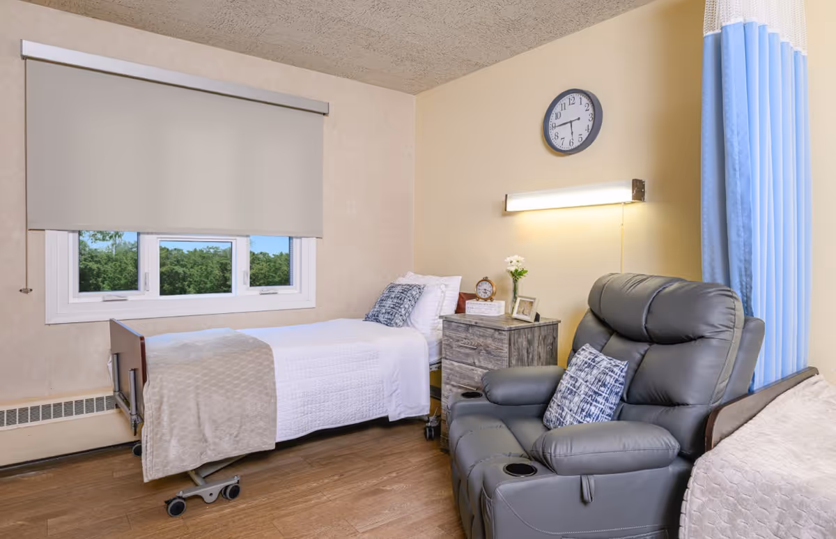 A small, neatly arranged bedroom in a senior living facility with a single hospital-style bed covered with white bedding and a beige blanket. Next to the bed is a wooden nightstand with a clock, a small vase with flowers, and a framed photo. A large gray recliner chair with a patterned pillow is positioned nearby. The room has a window with a beige roller shade partially down, a wall clock, and a blue privacy curtain on the right side.