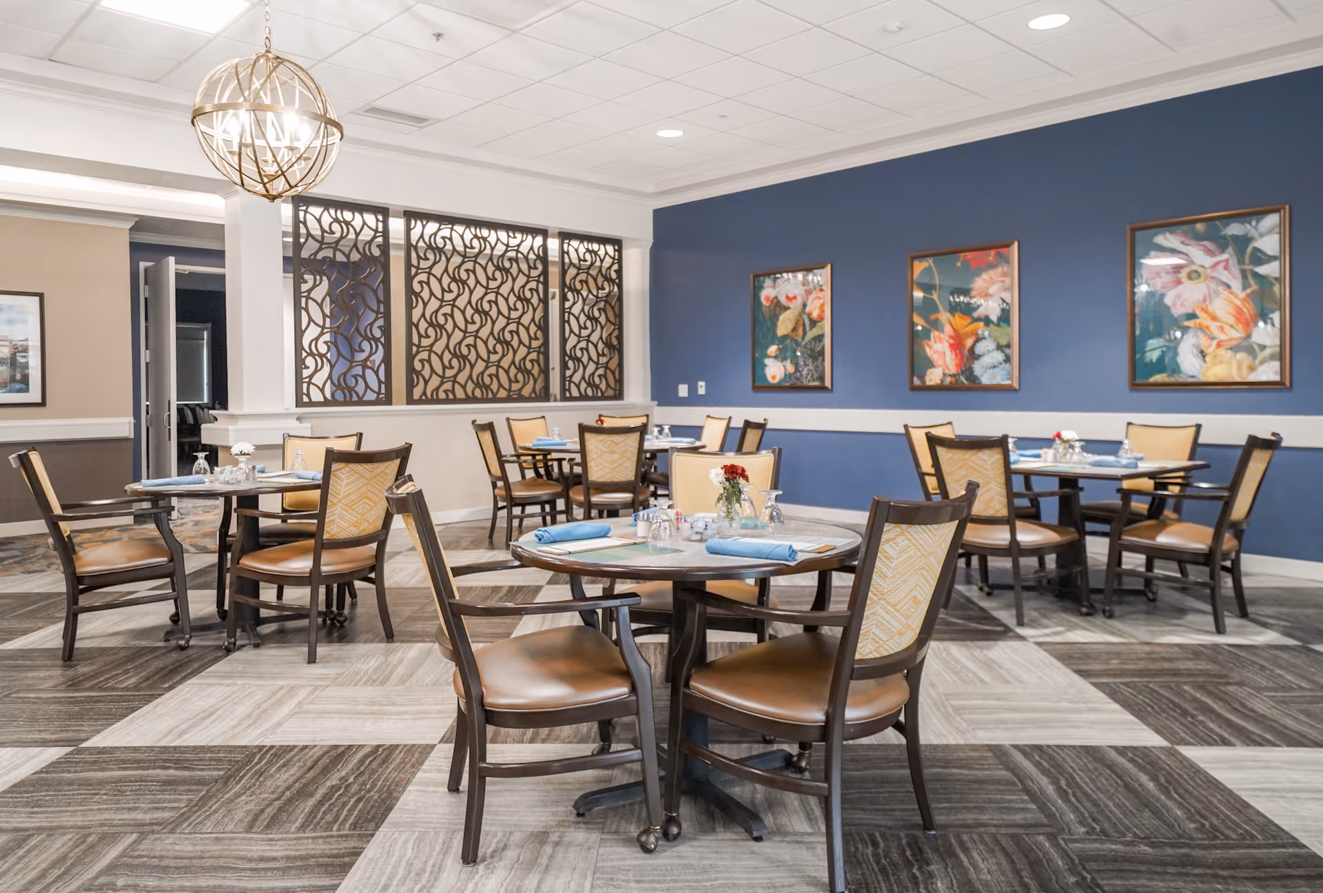 A dining room in a senior living facility with several round tables and chairs arranged neatly. Each table is set with blue napkins, glasses, and small flower vases. The room features a blue accent wall adorned with three framed floral paintings, a decorative metal partition, and a modern spherical chandelier hanging from the ceiling.
