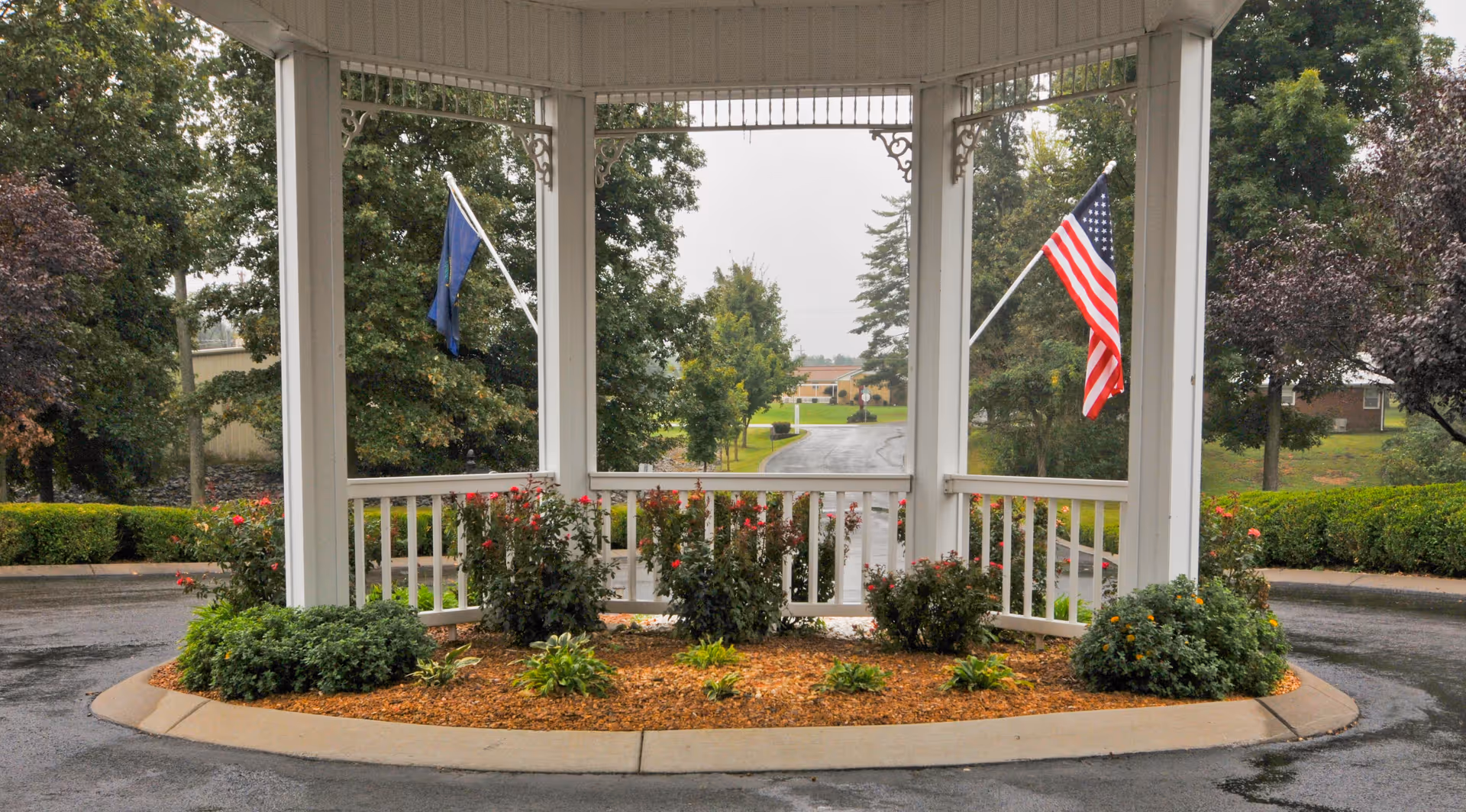 Covered entrance gazebo with landscaping, an American flag, and a driveway leading to the facility.