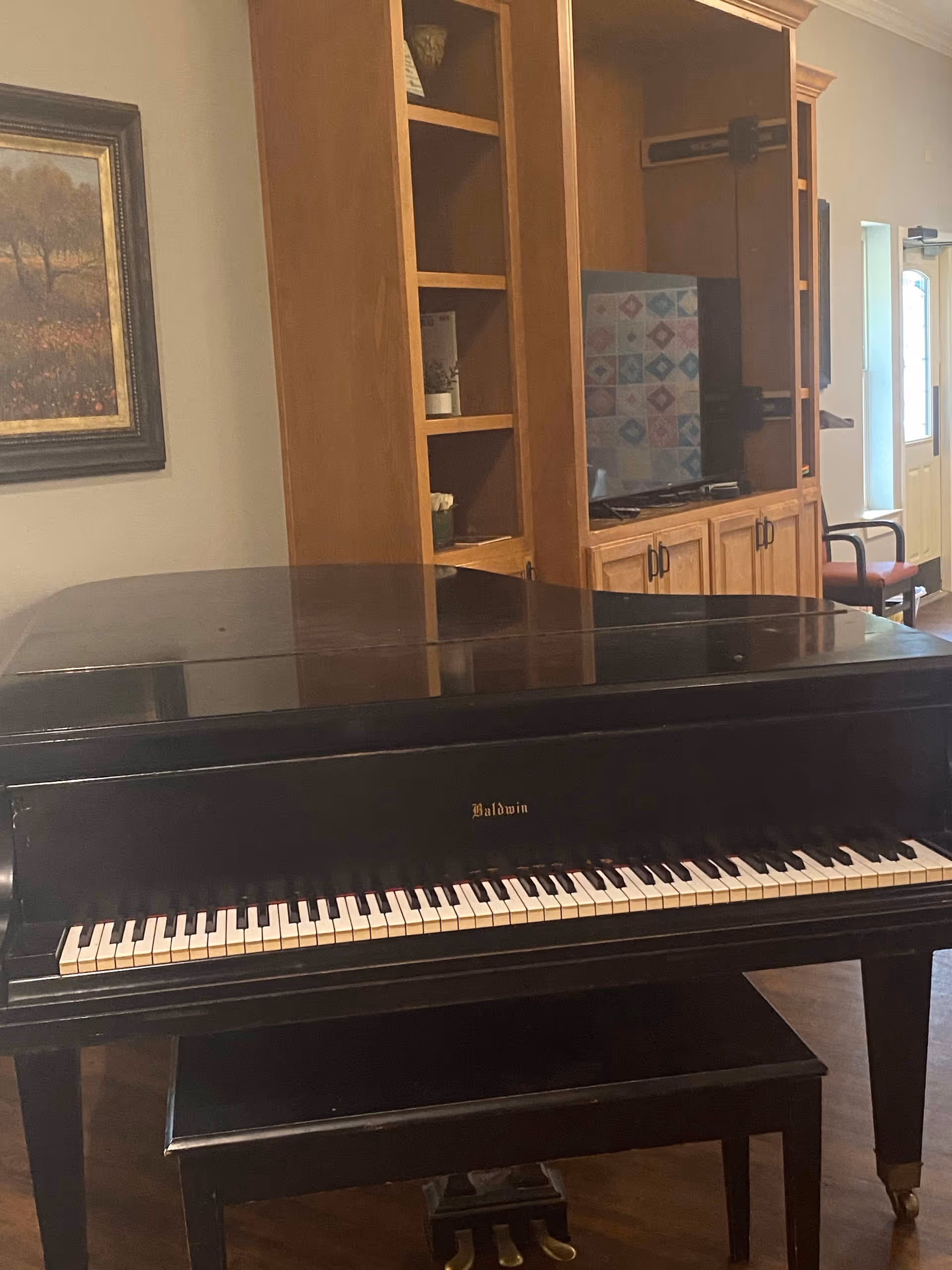 A black Baldwin grand piano with a matching bench in a room featuring wooden cabinetry with a mounted TV, a framed landscape painting on the wall, and a chair near a door with a window.