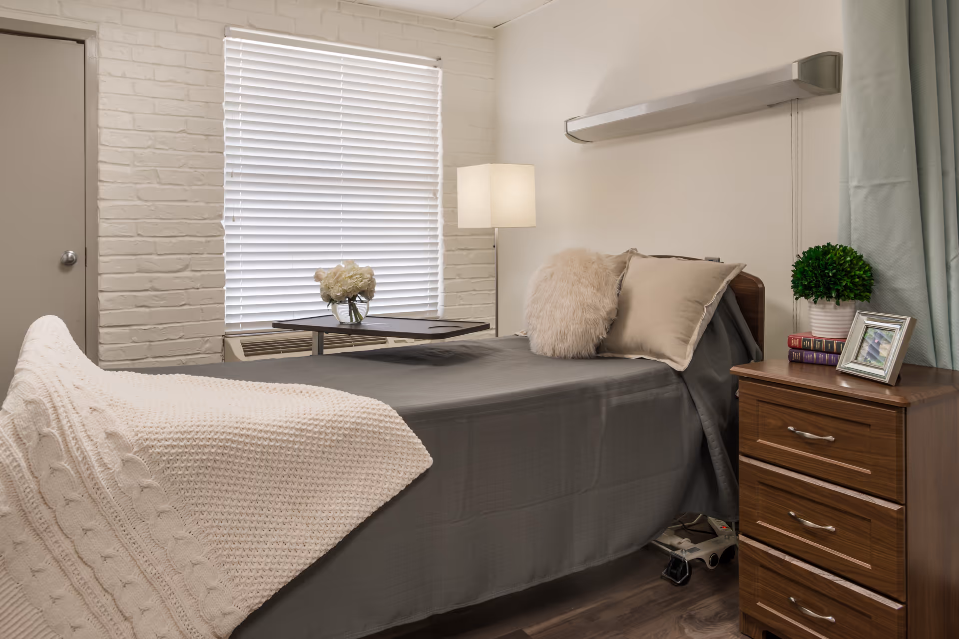 A neatly made single bed in a small bedroom with a nightstand, lamp, window blinds, and decorative pillows.