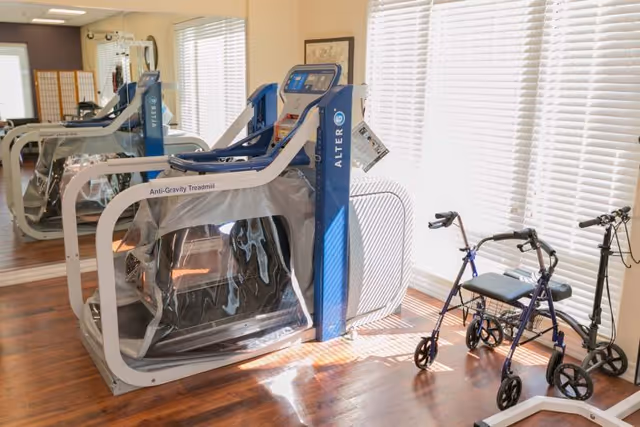 A bright room with wooden flooring featuring an Anti-Gravity Treadmill by AlterG covered in plastic, a blue walker with a seat, and a scooter near large windows with white blinds allowing sunlight to fill the space.