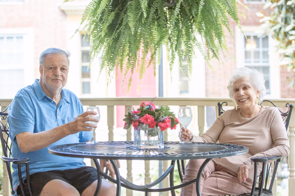 An elderly man and woman sitting at a round metal table on a porch, each holding a glass of water. There is a vase with pink flowers on the table and a large hanging fern above them. The background shows a brick building with windows and a red door.