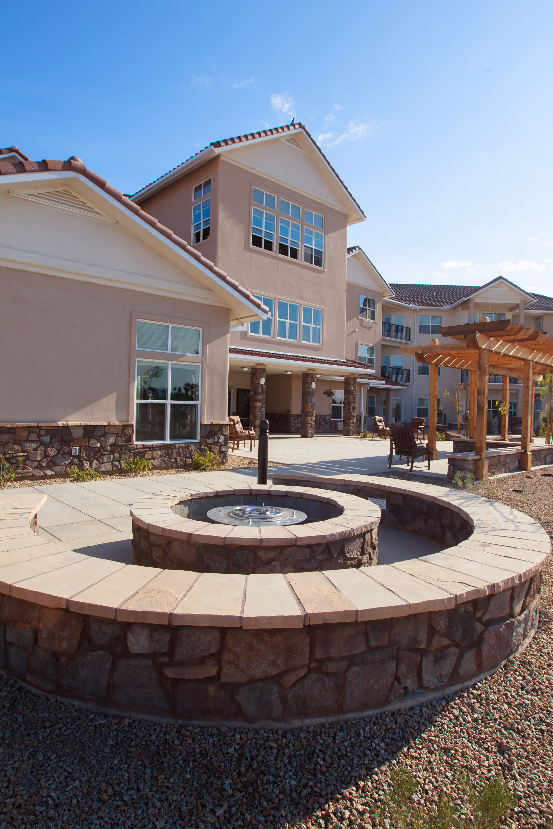 Outdoor seating area at a senior living facility with a circular stone fire pit in the foreground, surrounded by a stone bench. The building in the background has multiple windows and a covered patio area with chairs. There is also a wooden pergola to the right side of the image under a clear blue sky.