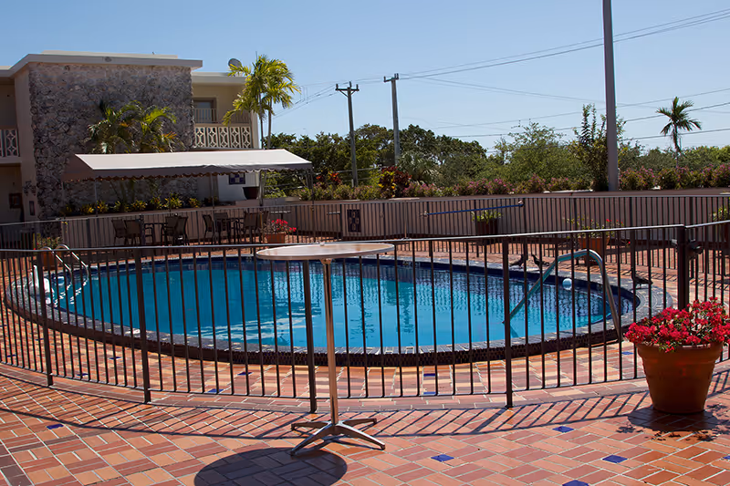Outdoor swimming pool area with a black safety fence around the pool, a round table in front of the fence, potted flowers on the tiled floor, and a building with stone and beige walls in the background under a clear blue sky.