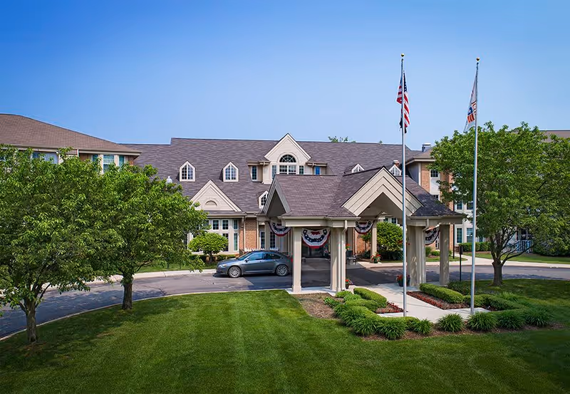 Front exterior view of Baldwin House Senior Living Oakland showing a large building with a covered entrance, two flagpoles with American flags, well-maintained green lawn, and trees on either side.