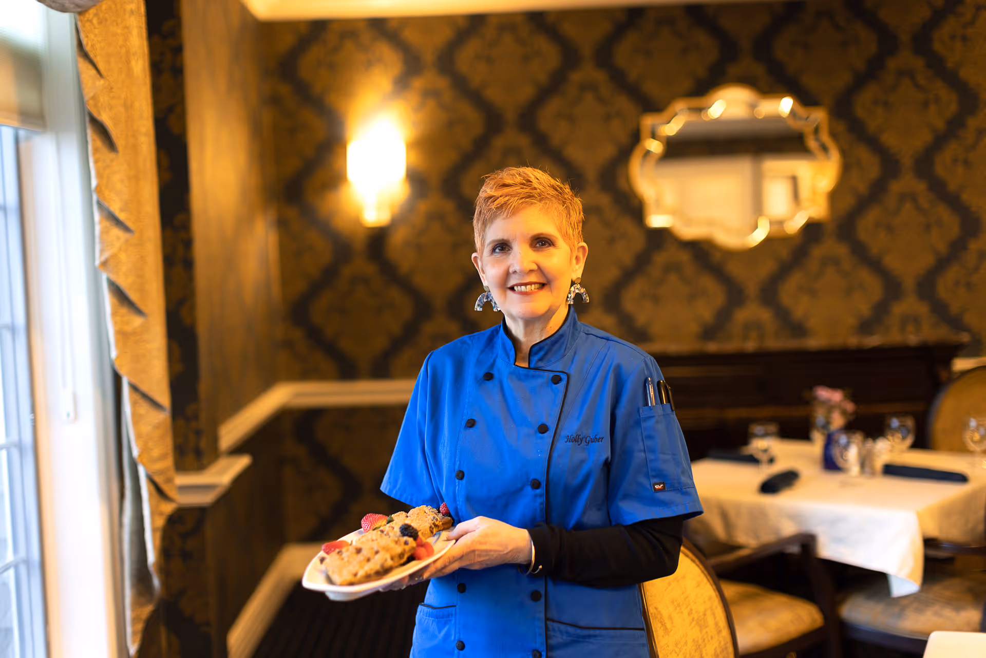 A woman in a blue chef's coat holding a plate of cookies with berries, standing in a dining room with elegant wallpaper, a decorative mirror, and tables set with glasses and napkins.