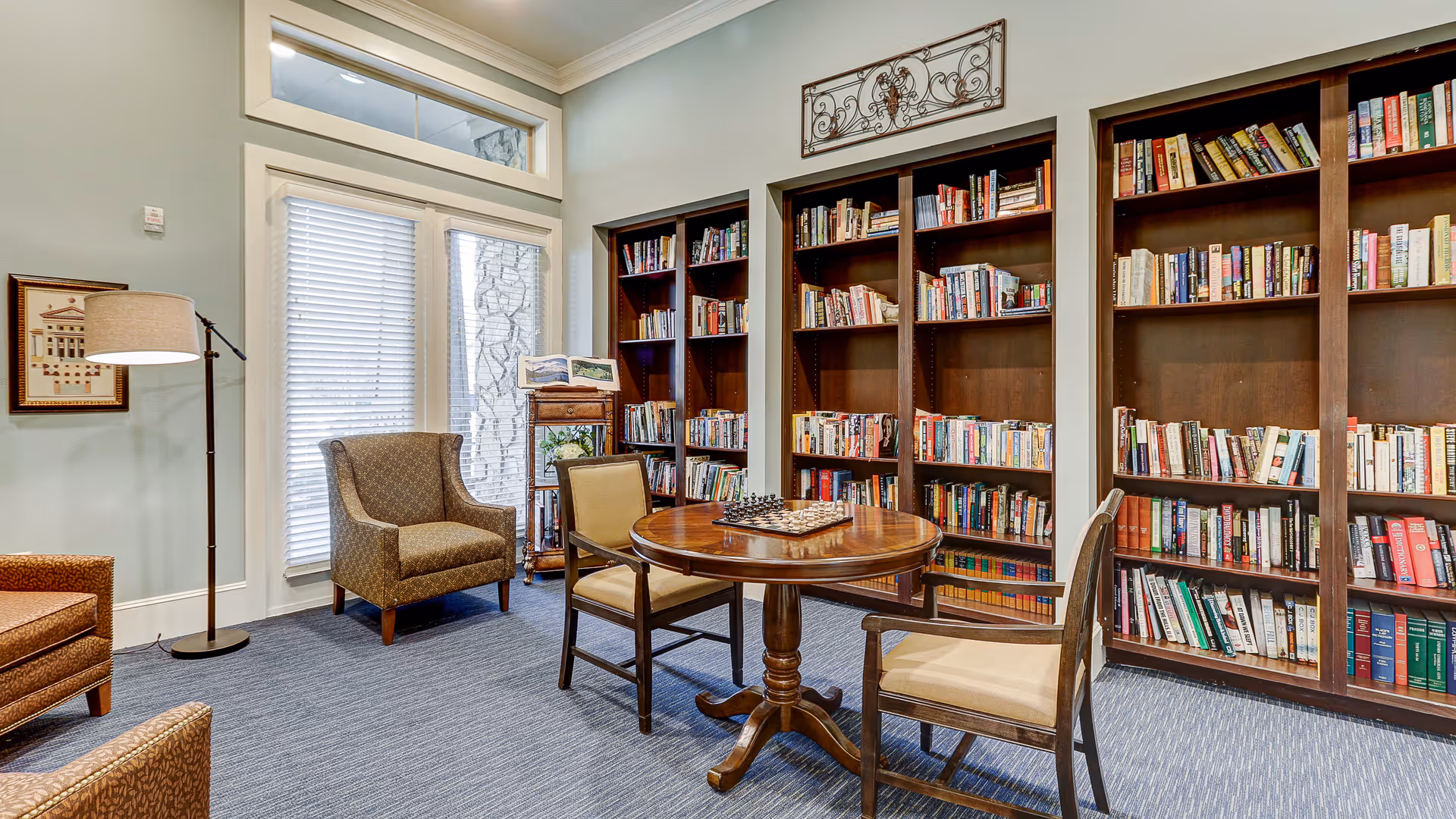 A cozy reading room with tall wooden bookshelves filled with books along the wall, a round wooden table with a chessboard on it, and four chairs around the table. There is a floor lamp and a framed picture on the wall, with large windows covered by blinds letting in natural light.