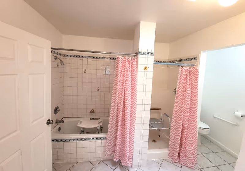 Bathroom with two separate bathing areas divided by a tiled column. The left side has a bathtub with a white shower chair inside and a pink patterned shower curtain partially drawn. The right side has a walk-in shower with a similar white shower chair and matching pink patterned shower curtain. To the right, there is a toilet with a grab bar and a toilet paper holder on the wall. The floor is tiled and the walls are white.
