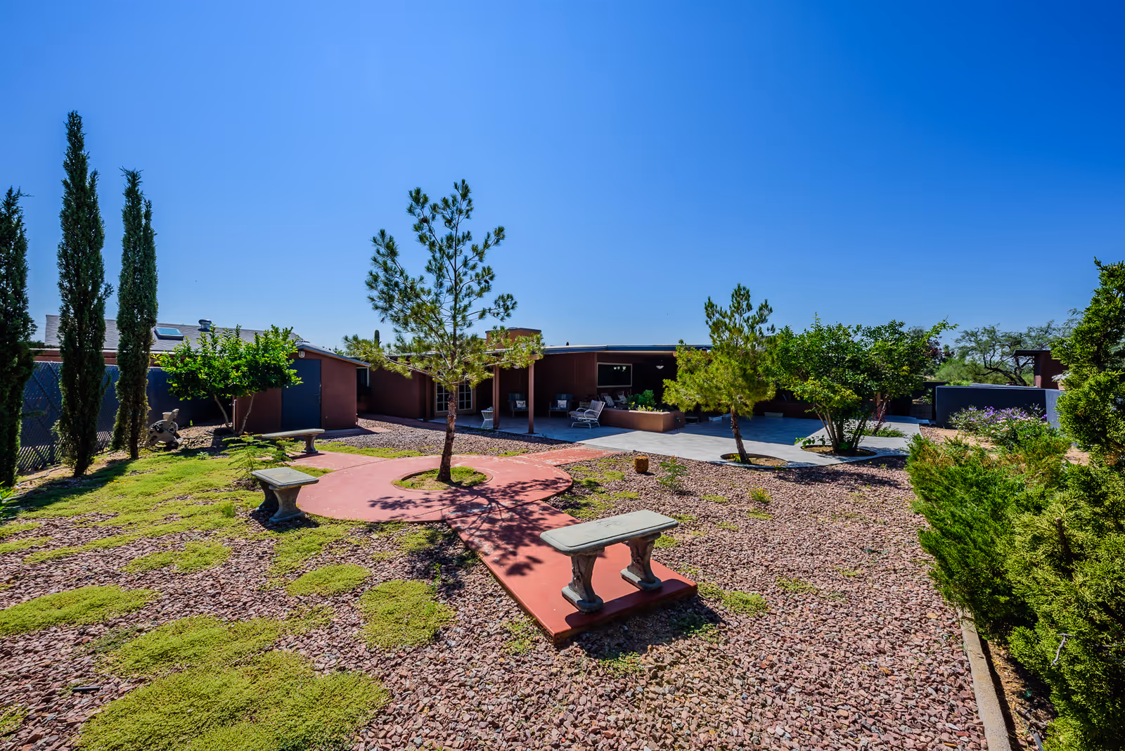 Sunny courtyard with gravel landscaping, scattered trees and concrete benches in front of a single-story assisted living building.