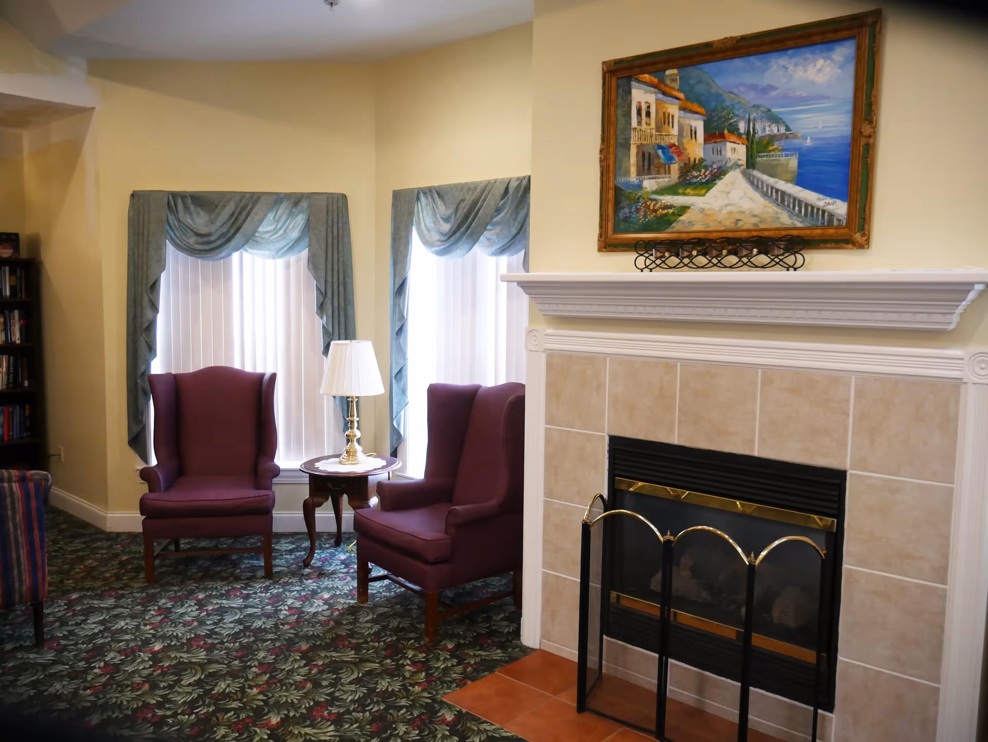 A cozy seating area with two purple wingback chairs beside a side table and lamp facing a tiled fireplace topped by a coastal painting.